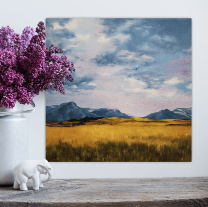 Christina Gouldsborough original oil painting of a wheat field with mountains in the background and lilac hued sky, on a white wall behind a shelf and vase with lilacs