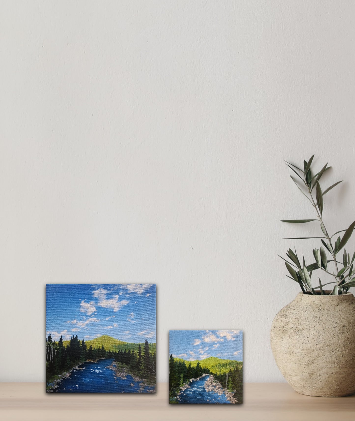 Two small 6 inch by 6 inch and 4 by 4 inch oil paintings of a river surrounded by trees and miles of forest, beside a plant on a wood shelf