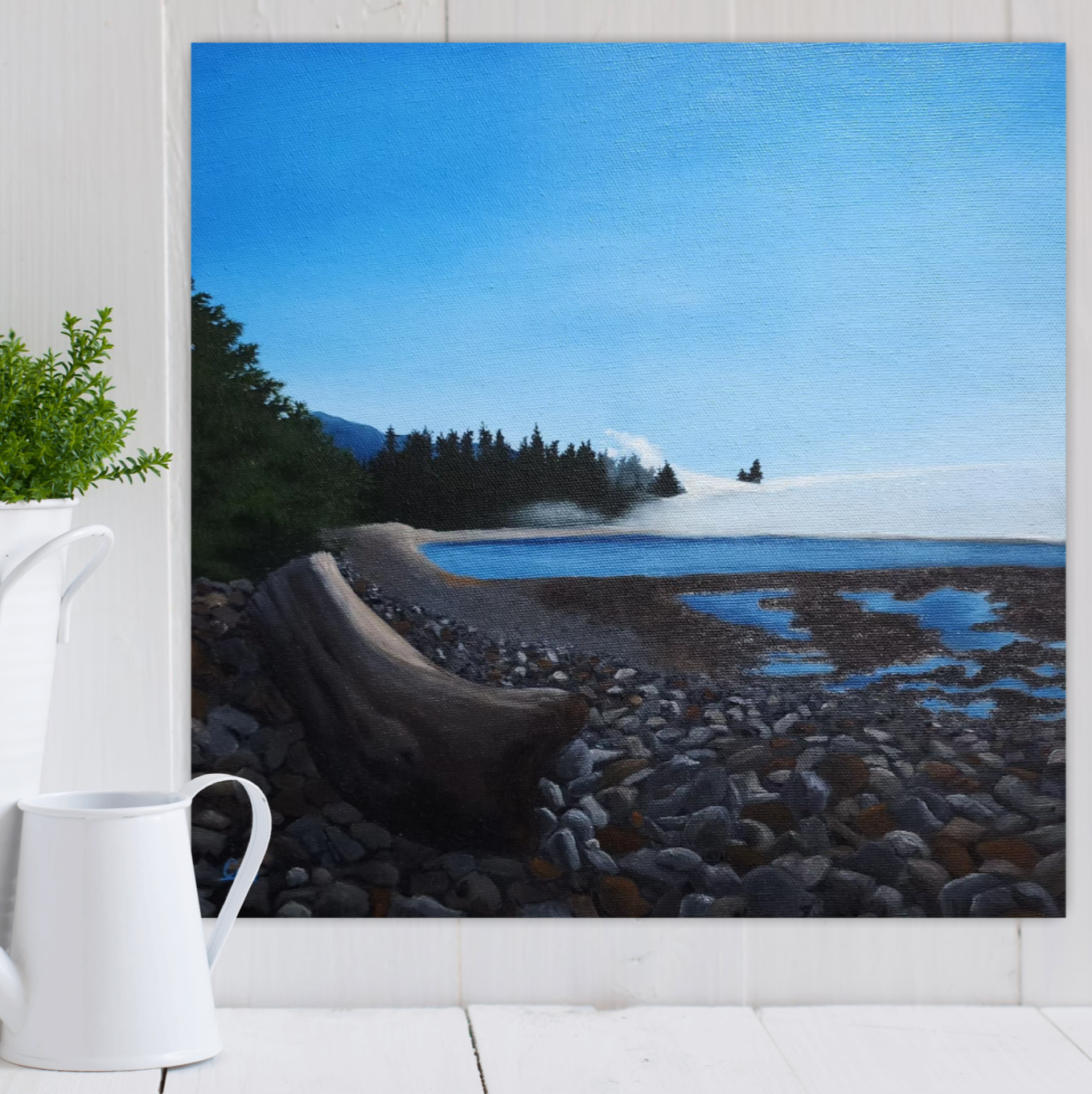 Christina Gouldsborough's original landscape painting "Misty Driftwood Daydream" of a Vancouver rocky shore with a large piece of driftwood, on a white wall, with a plant and water can in front on a table