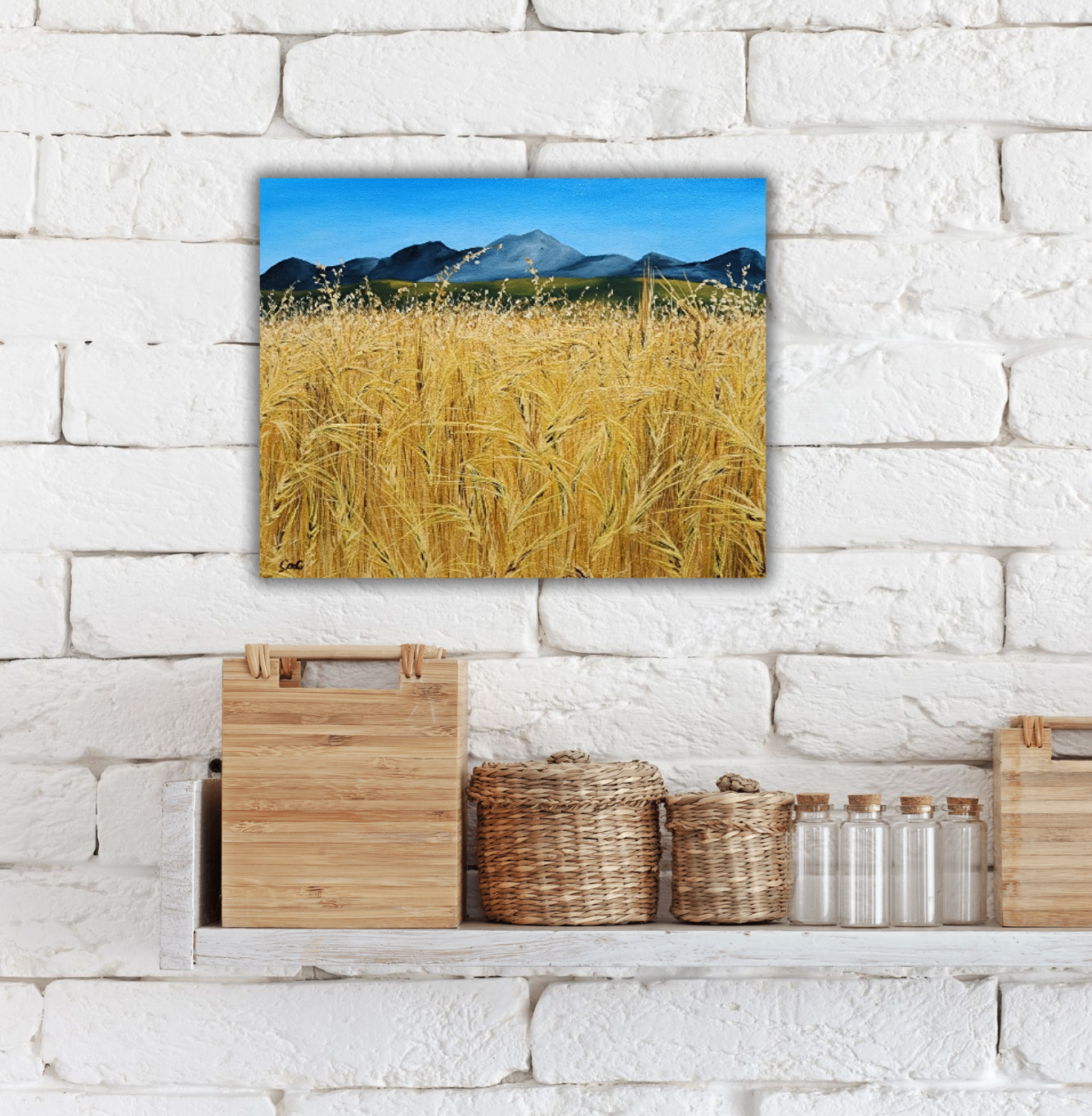 Christina Gouldsborough original oil painting "Picnic In the Wheat" of a up close wheat field with distant mountains in the distance on a shelf behind some wood and wicker decorations