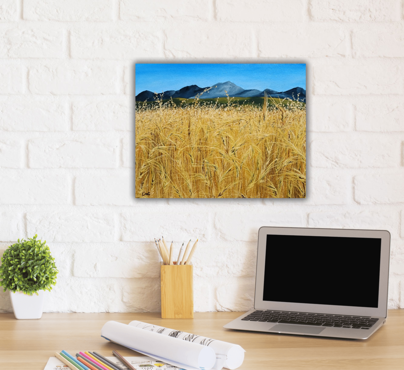 Christina Gouldsborough original oil painting "Picnic In the Wheat" of a up close wheat field with distant mountains in the distance. on a desk behind a laptop plant, pencils and papers