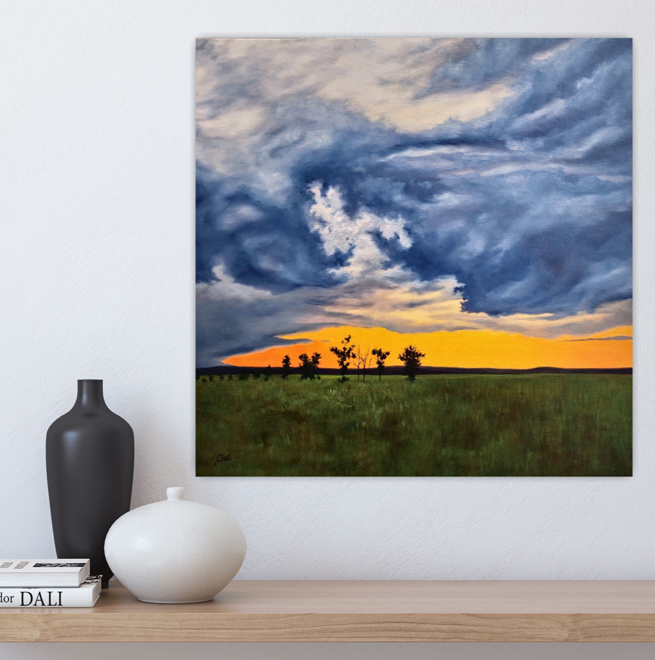 Evening Dichotomy - 16"x16" oil on canvas landscape painting of a prairie field with a few trees and storm clouds at sunset on a white wall behind a wood shelf