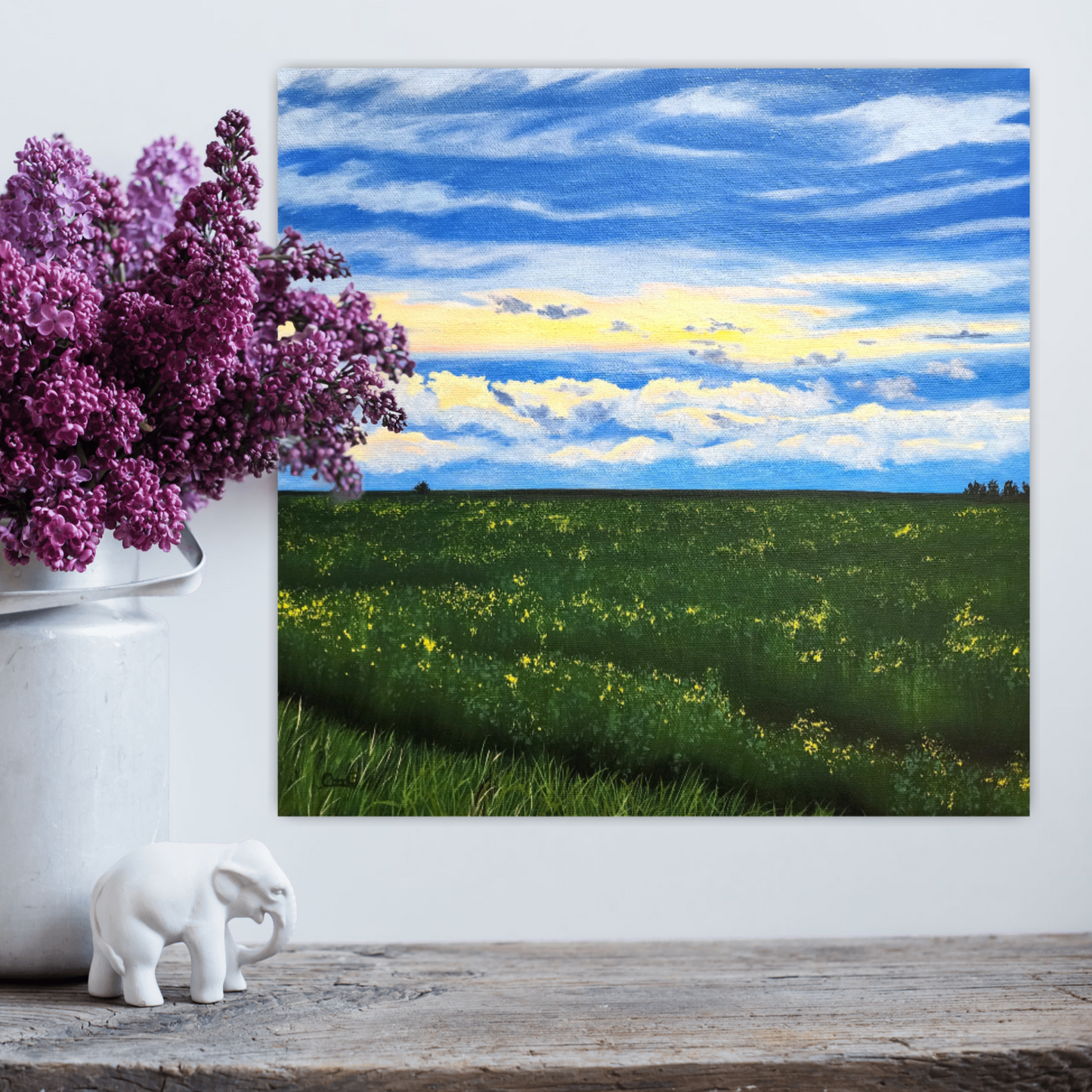 After the Rain Canadian landscape painting of a summer canola field after a rain storm by Christina Gouldsborough on a white wall behind purple flowers